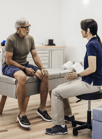 The image is of a man sitting on a physiotherapy treatment table. His physiotherapist is sitting on a stool talking to him. 